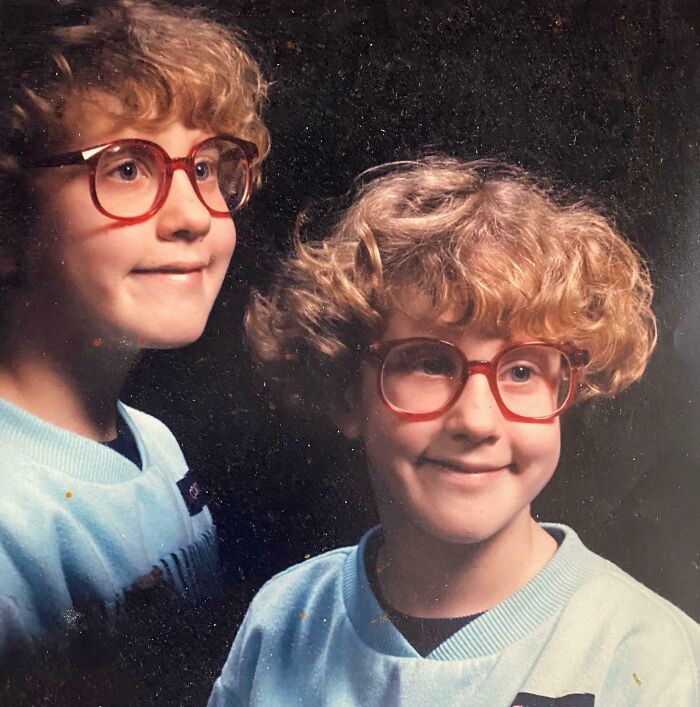 Childhood photo of a smiling boy with red glasses and curly hair wearing a light blue sweatshirt in a studio portrait.