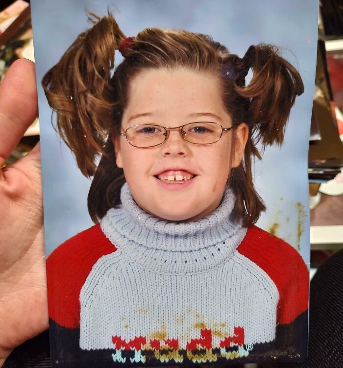 Childhood photo of a girl with glasses and pigtails wearing a colorful sweater, showcasing an awkward and funny moment.