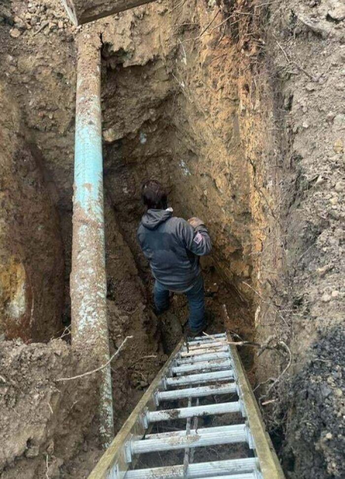 Worker standing in a deep trench next to a pipe without proper safety measures, ignoring safety protocol hazards.