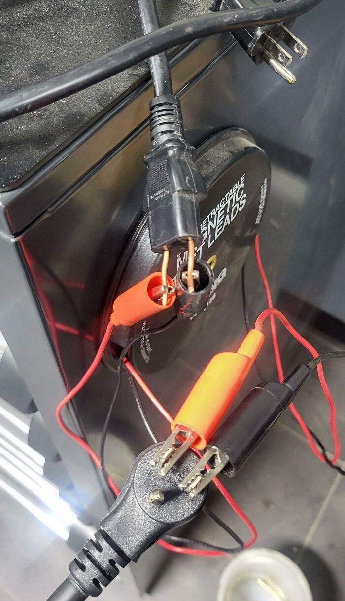 Exposed electrical wires connected to power plugs with testing leads, showing employees blatantly ignoring safety protocol hazards.