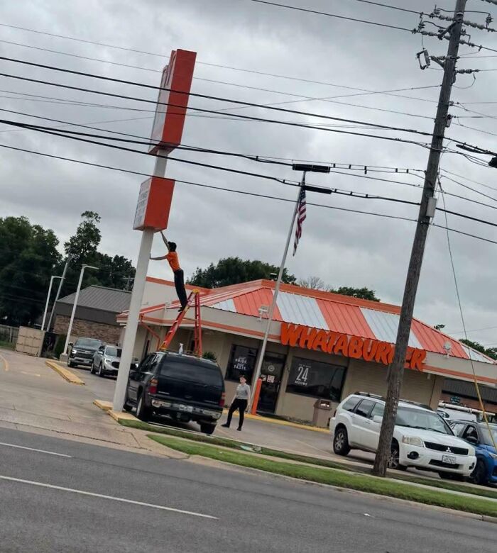 Worker blatantly ignoring safety protocol by standing on a ladder on a truck to reach a high sign near power lines.