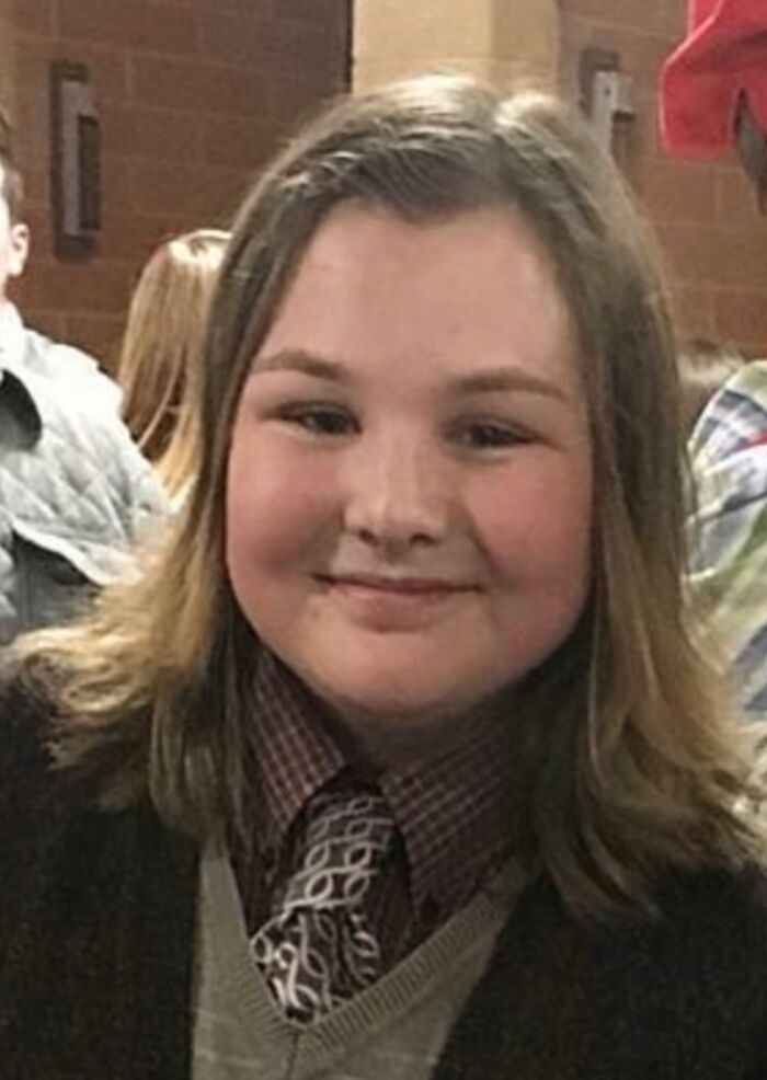 Young girl smiling indoors at an event, wearing a patterned tie and sweater, part of hilariously awkward childhood photos.
