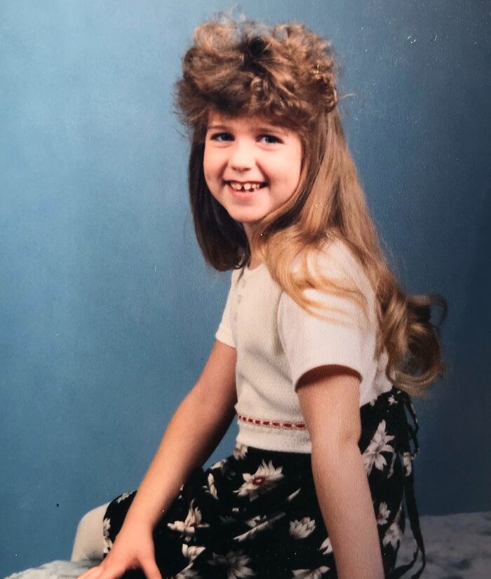 Young girl with a voluminous hairstyle and floral skirt, smiling awkwardly in a vintage childhood photo.