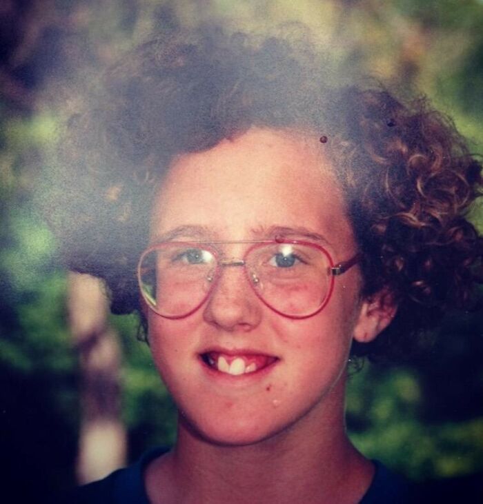 Child with curly hair and large red glasses smiles awkwardly in a hilariously awkward childhood photo.