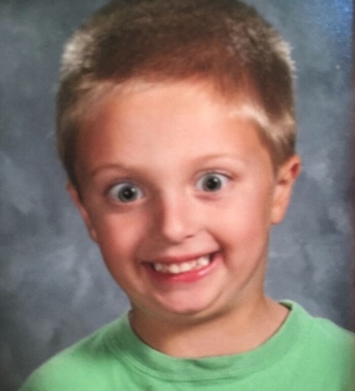 Young boy making a hilariously awkward face in a childhood photo with a green shirt and a gray background.