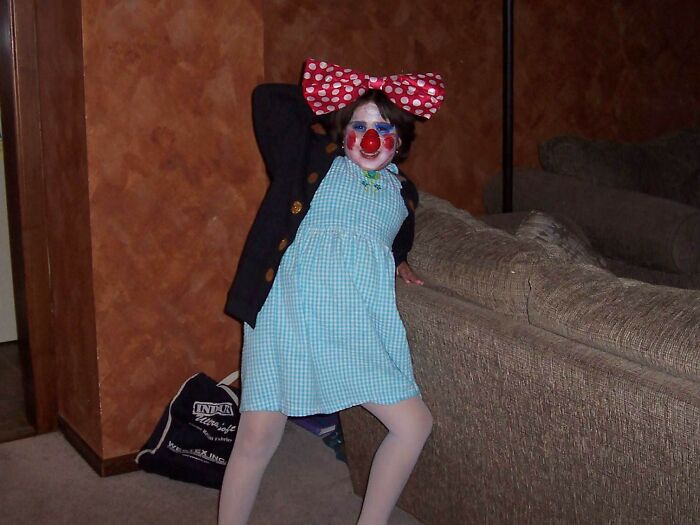 Child in awkward childhood photo wearing clown makeup, large bow, and blue dress striking a playful pose indoors.