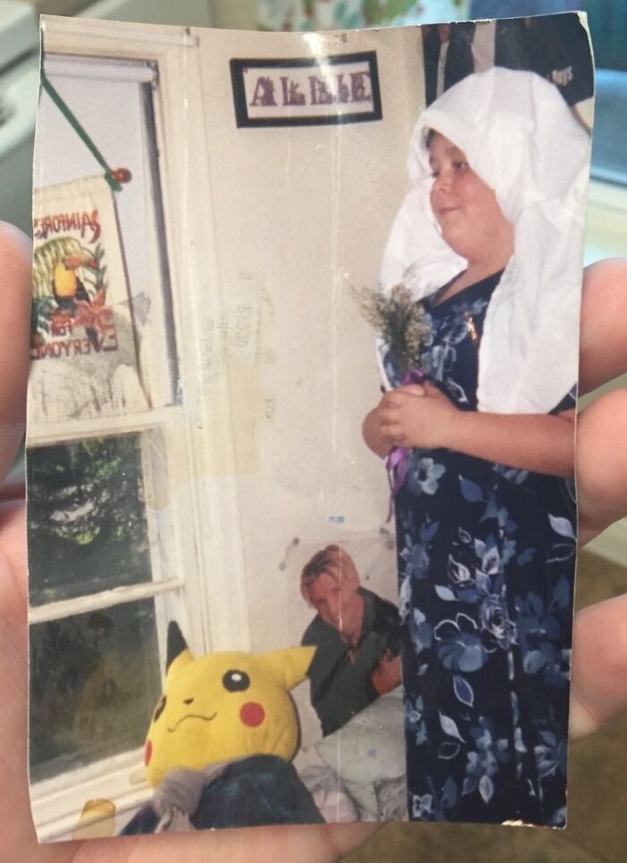 Child wearing a floral dress and white headscarf holding flowers in an awkward childhood photo with Pikachu plush.