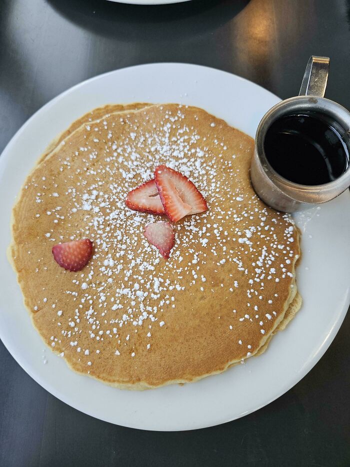 Outrageous meal of a single pancake lightly dusted with powdered sugar and a few strawberry slices, served with syrup.