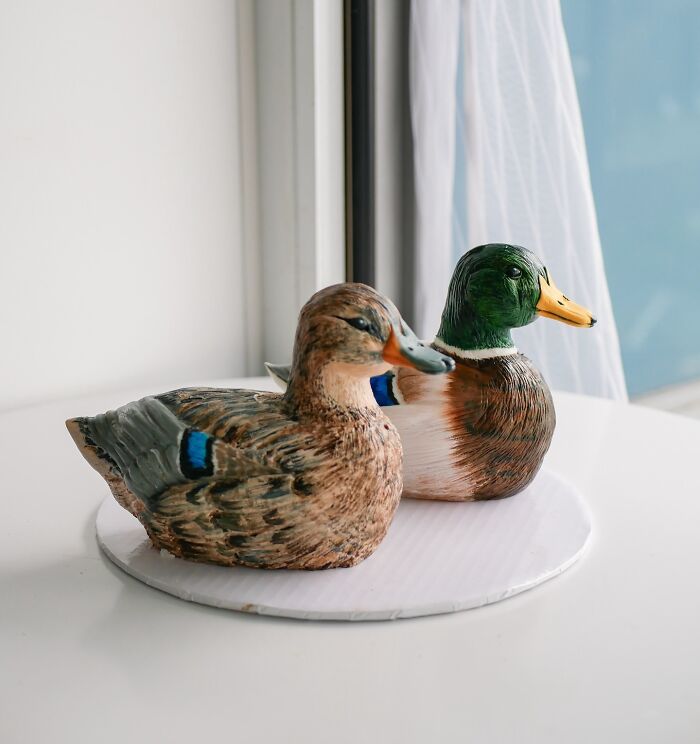 Two masterfully crafted duck-shaped cakes showcasing creativity and detailed edible art on a white surface near a window.