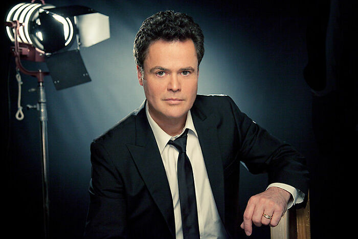 Donny Osmond wearing a black suit and tie, seated in a studio setting with professional lighting in the background.