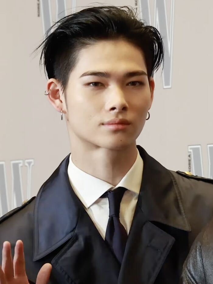 Ni-Ki wearing a black trench coat and tie, posing at an event with earrings and styled hair.