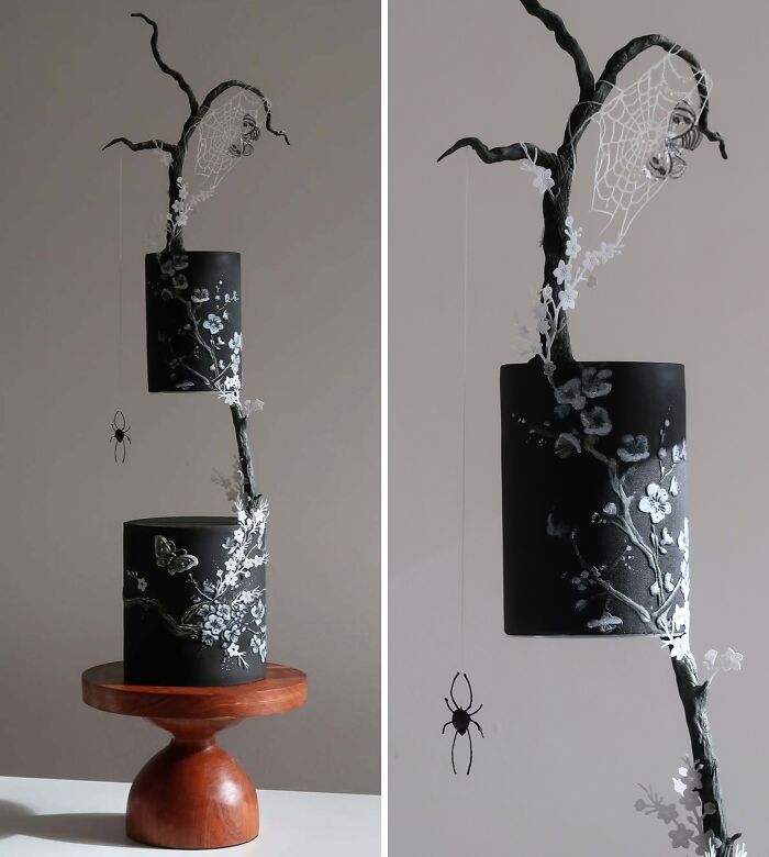 Black two-tier cake with white floral designs and a spider web, featuring a hanging spider, showcasing masterfully crafted cake art.