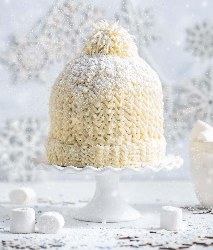 Cake shaped and decorated like a knitted winter hat with white frosting and powdered sugar on a cake stand