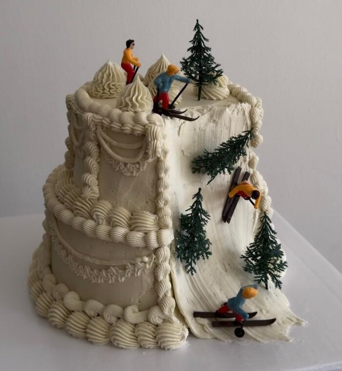 White creatively crafted cake designed as a snowy ski slope with miniature skier figurines and pine trees.