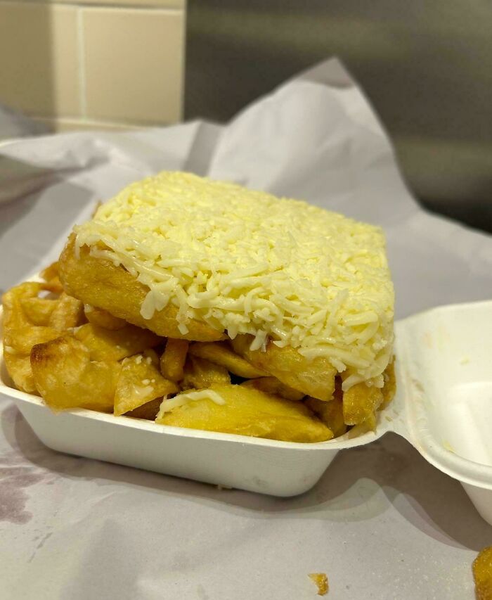 Takeout container with an unusual meal of fries topped with a fried square covered in shredded cheese, an outrageous meal fail.