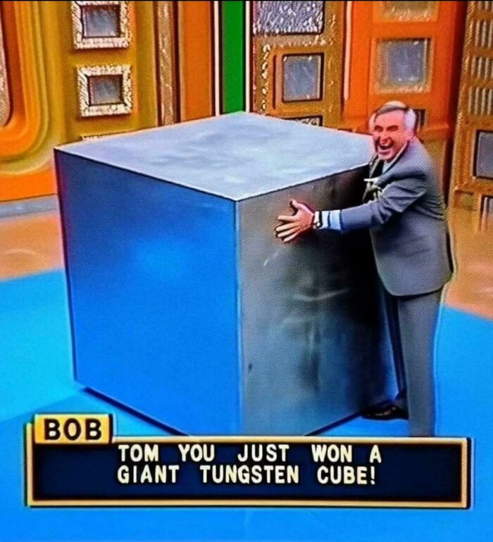 Man in gray suit excitedly hugging a giant tungsten cube on a colorful game show stage solving weird math problems.