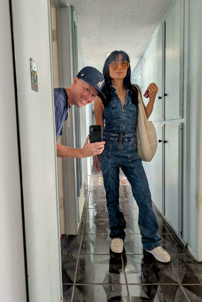Two people posing in a hallway mirror selfie wearing casual outfits showcasing cool secondhand store finds.
