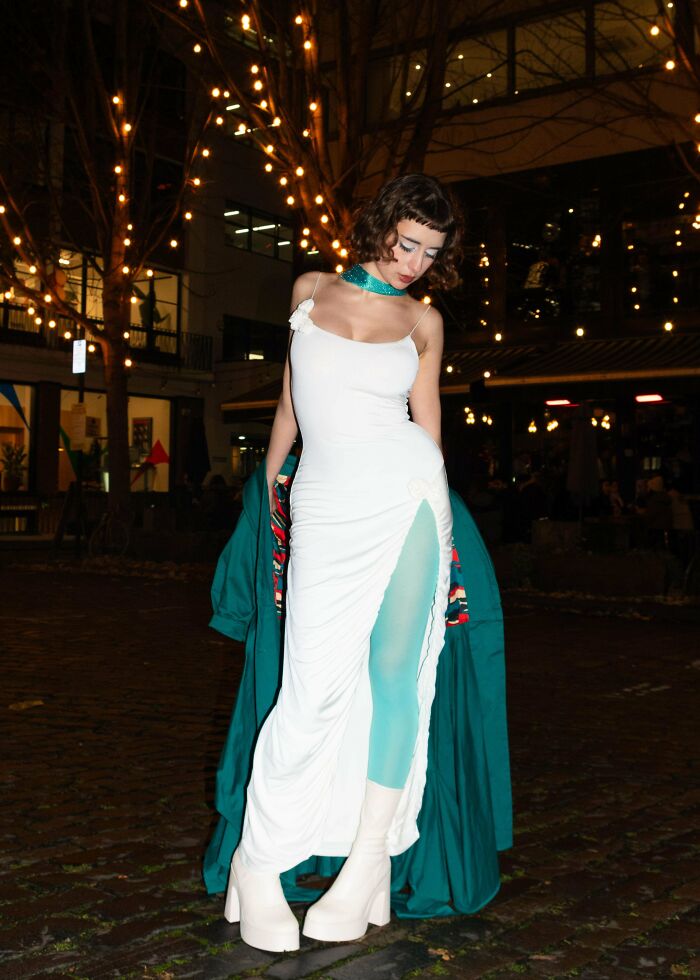 Woman wearing a stylish secondhand outfit with a white dress and teal tights under warm glowing outdoor lights at night.