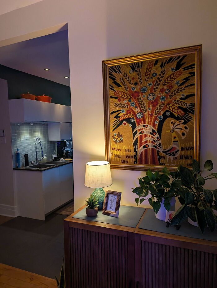 Cozy home corner with secondhand store art tapestry, wooden cabinet, table lamp, and green plants adding vintage charm.