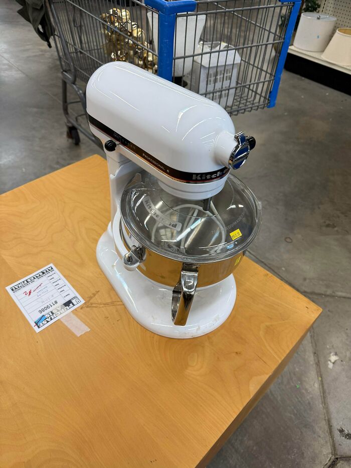 White KitchenAid stand mixer on wooden table in secondhand store with shopping cart in background and price tag visible