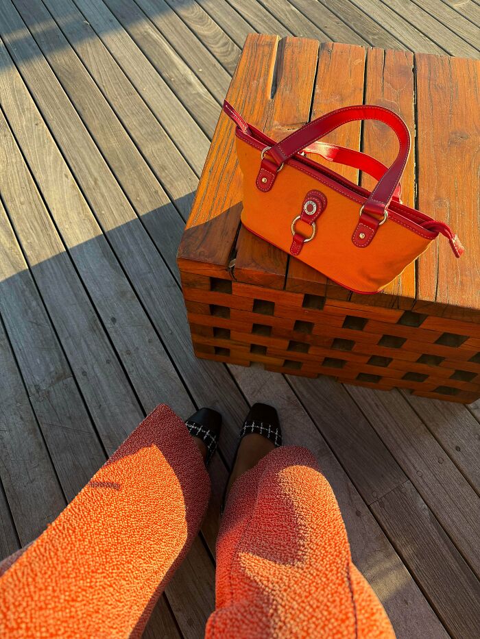 Orange handbag placed on wooden table with person wearing orange coat and black shoes nearby, reflecting secondhand store finds.