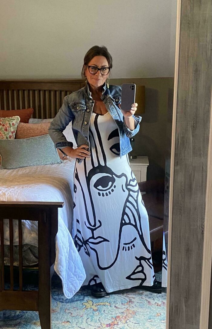 Woman taking mirror selfie wearing a secondhand store dress with abstract face print and a denim jacket in a bedroom.