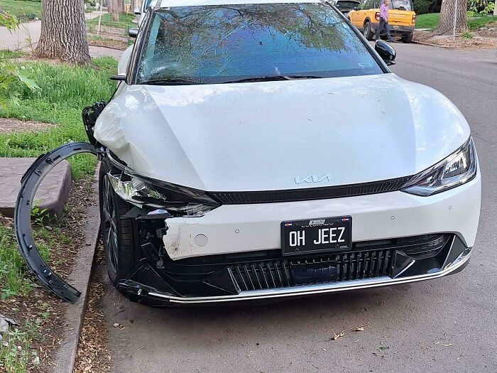 White car with damaged front bumper and personalized license plate reading OH JEEZ parked on a residential street.