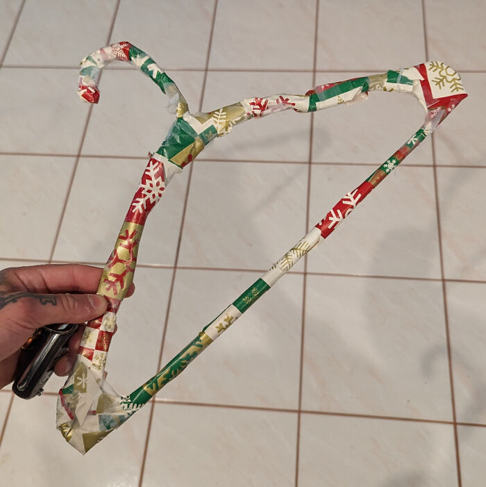 Hand holding a hanger creatively wrapped in funny Christmas gift wrap with colorful holiday patterns on tile floor.