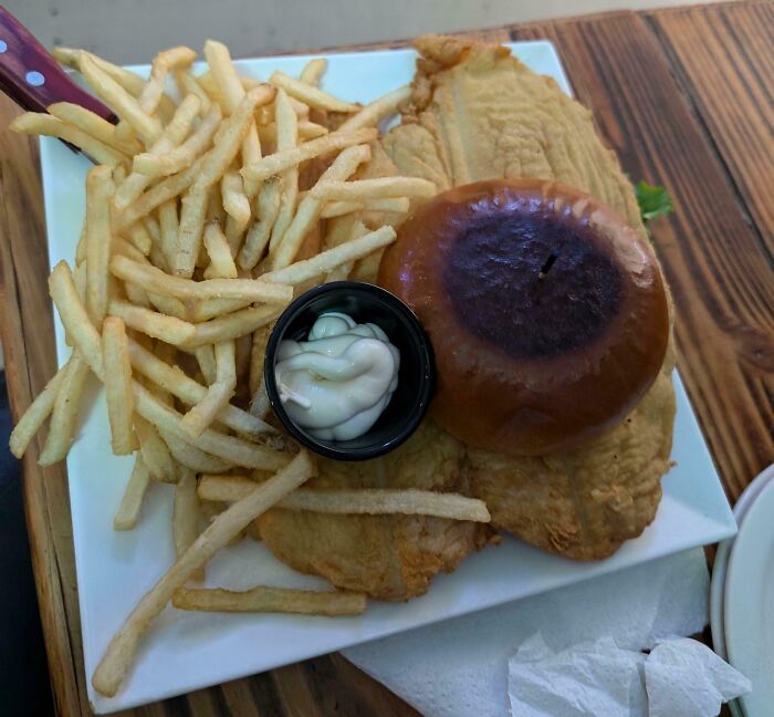 Plate of food showing an outrageous meal with burnt sandwich bun, thin fries, and a small container of sauce on a wooden table.