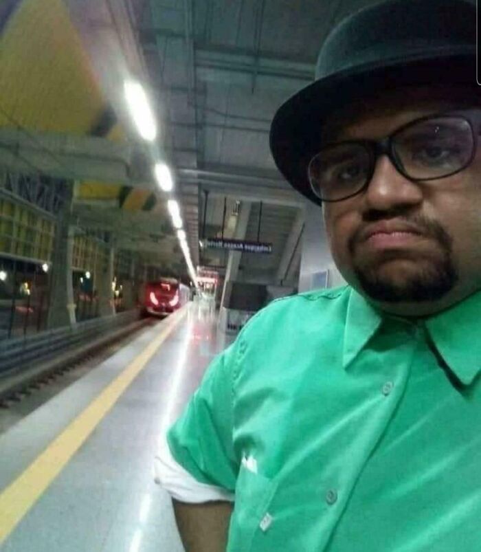 Man in a green shirt and black hat taking a selfie on a train platform with a train approaching in the background.