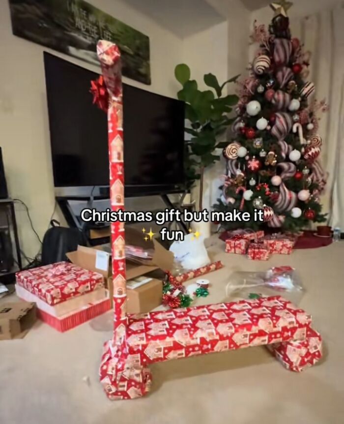 Humorous gift wrap creating a scooter-shaped Christmas present in a festive living room with a decorated tree.