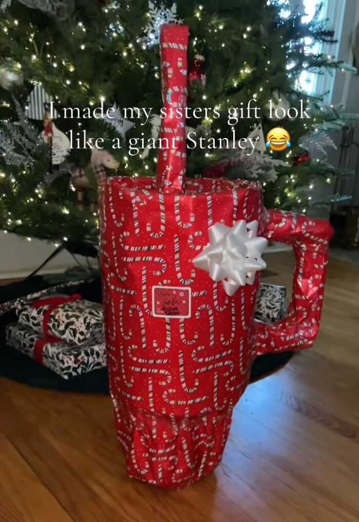 Funny Christmas gift wrap designed as a giant Stanley cup, decorated with candy cane print and a white bow by Christmas tree.
