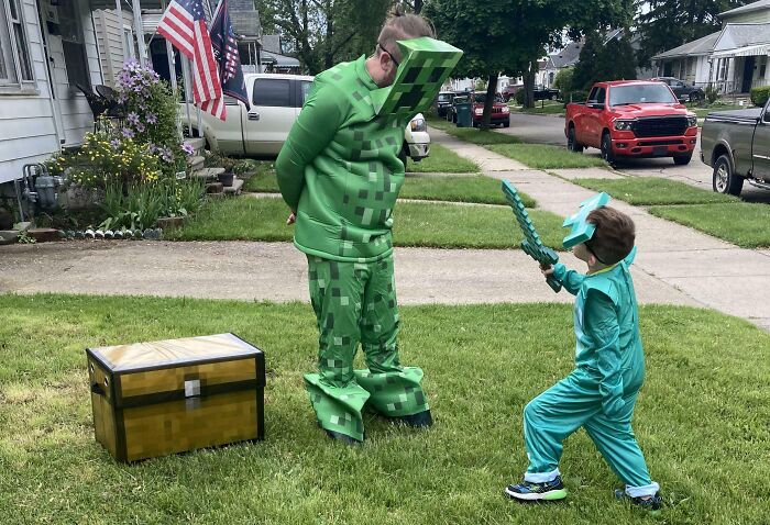 Dad and child dressed in green Minecraft costumes playing outside with a toy sword and chest on the grass.