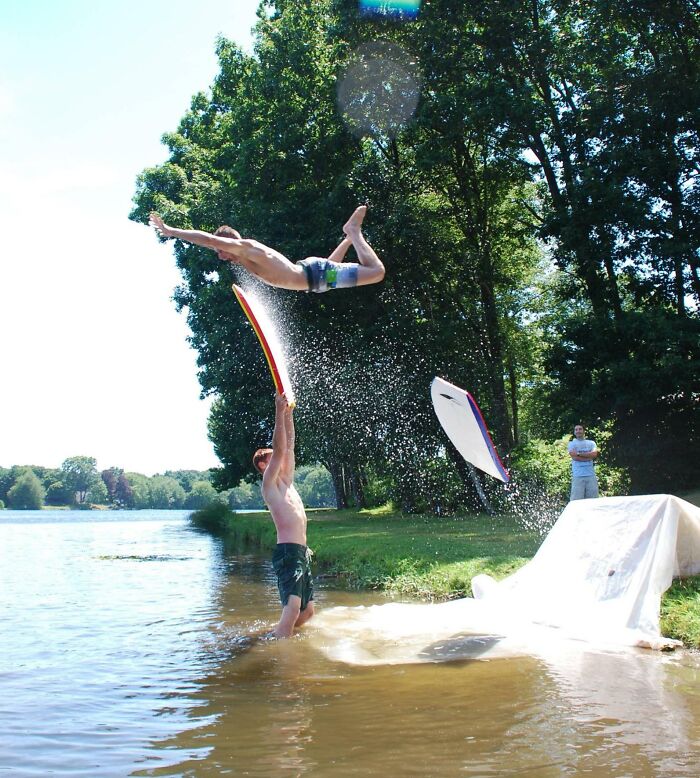 Two men by a lake captured at the perfect time as one flips midair with water splashing during a funny and interesting moment.
