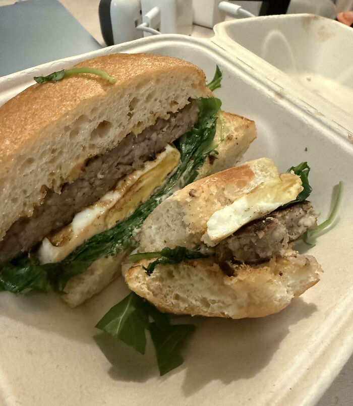 Outrageous meal showing a small burger with uneven ingredients and missing expected toppings in a takeout container.