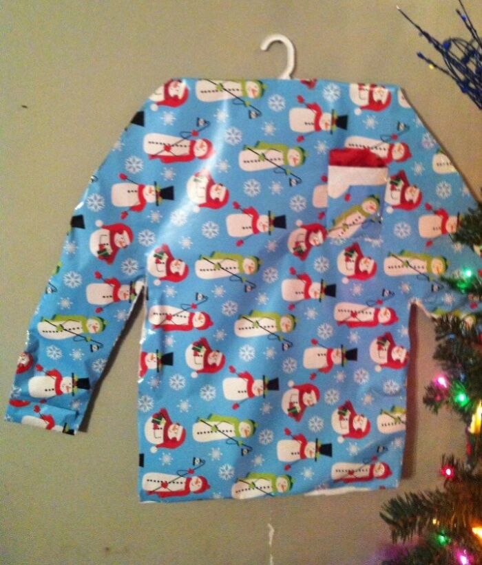 Sweater wrapped in funny Christmas gift wrap with snowmen on a wall near a decorated holiday tree.