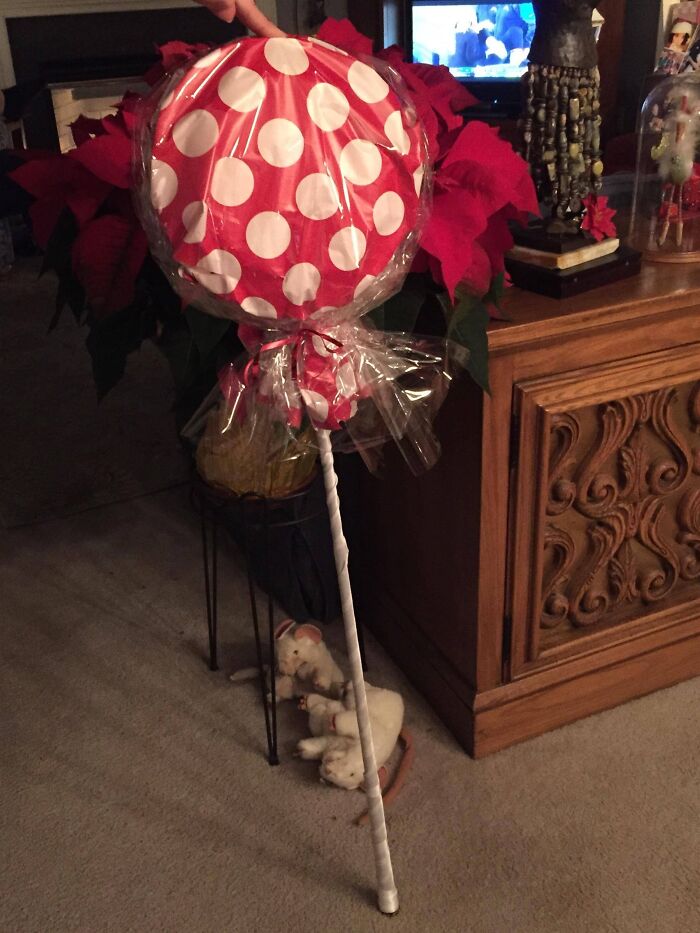 Large gift wrap shaped like a red and white polka dot lollipop with a stick, placed near poinsettias and holiday decor.