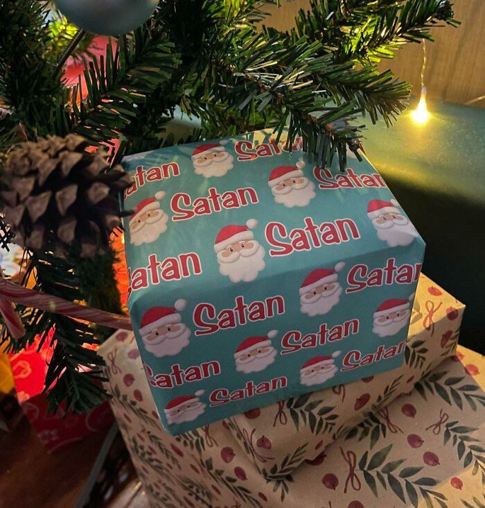 Christmas gift wrapped in humorous paper with Santa faces and the word Satan under a decorated tree with lights and pinecones.