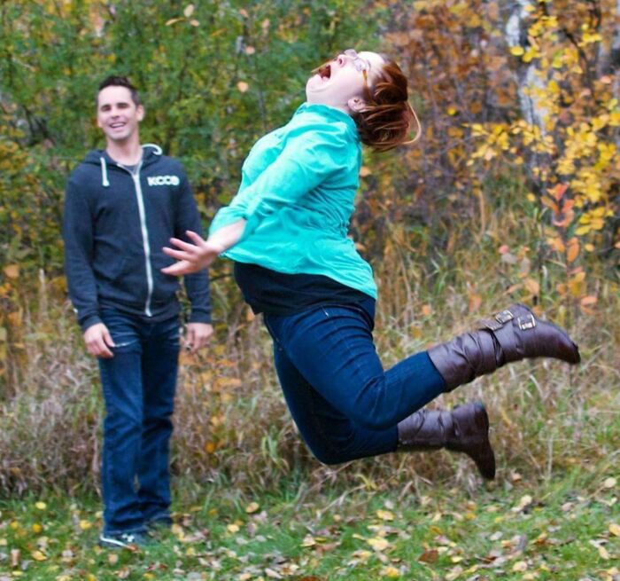 Woman mid-air in an awkward pose while jumping outdoors, with a man smiling in the background in a funny, interesting, weird pic.