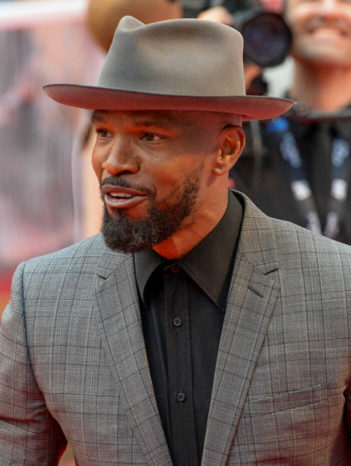 Jamie Foxx wearing a gray hat and checkered suit jacket at a public event, highlighting his bio and career achievements.