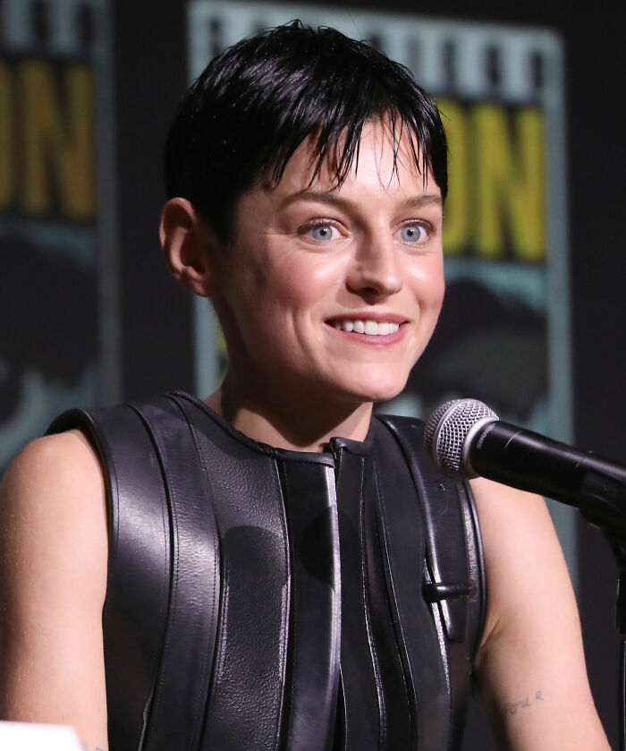 Emma Corrin speaking at a panel, wearing a black leather outfit with short hair and a bright expression.
