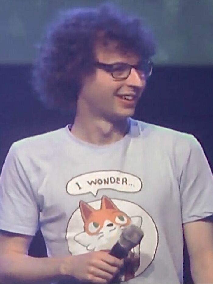 Joseph Garrett smiling on stage, wearing glasses and a cartoon fox t-shirt, holding a microphone during a talk.
