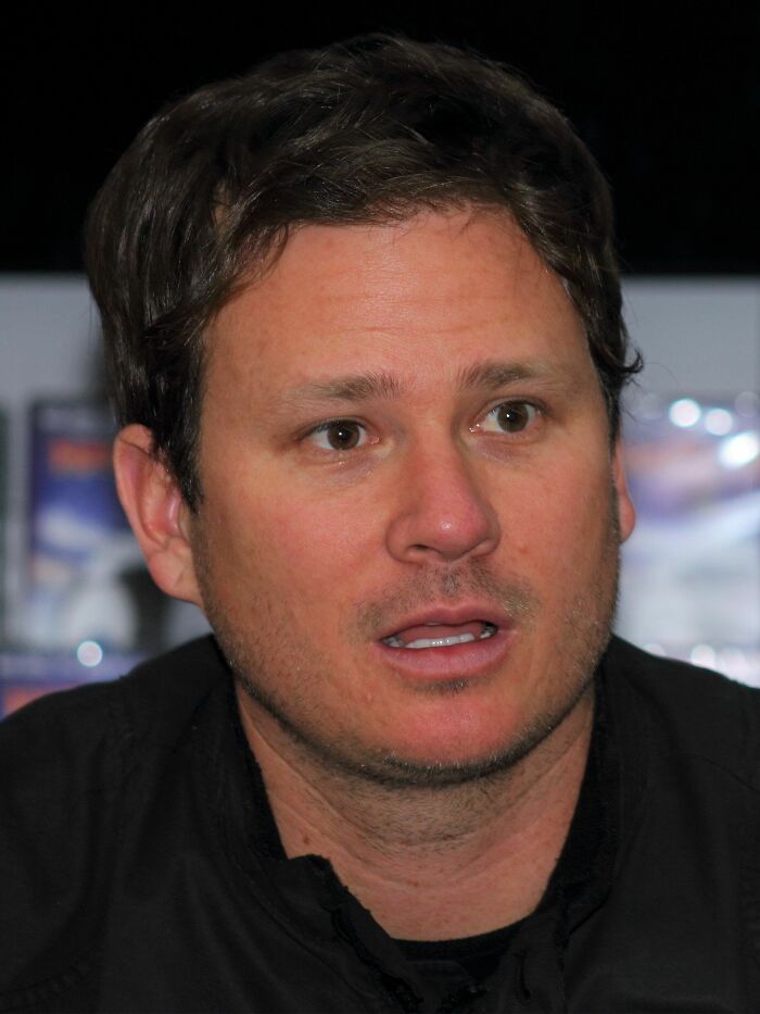 Tom DeLonge speaking during an interview, wearing a black jacket with a blurred background and focused expression.
