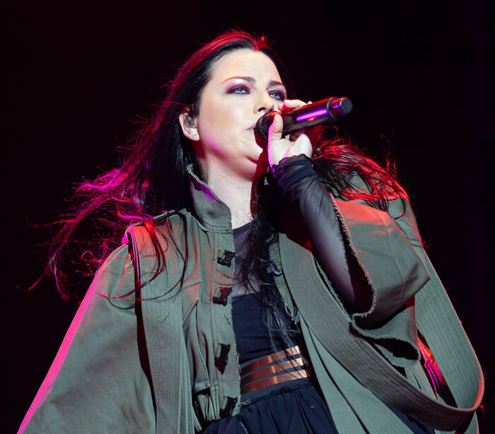 Amy Lee performing on stage, singing into a microphone with dynamic lighting and flowing dark hair.