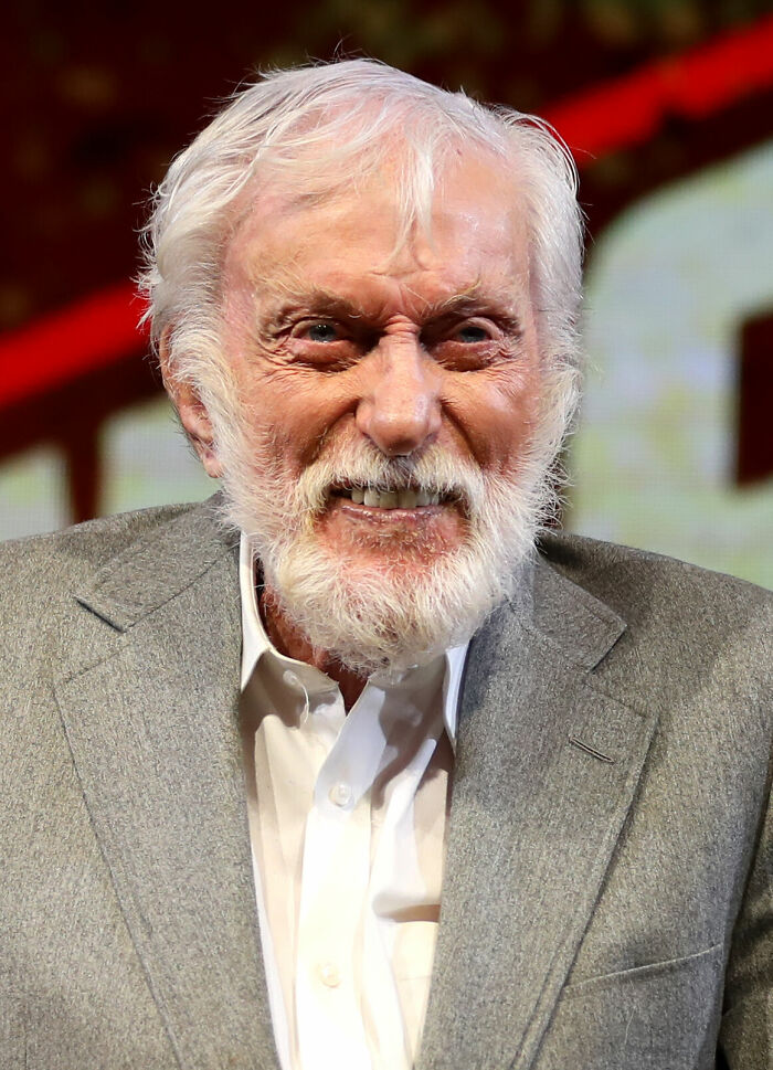 D**k Van D**e smiling with white hair and beard, wearing a gray suit jacket and white shirt at an event.