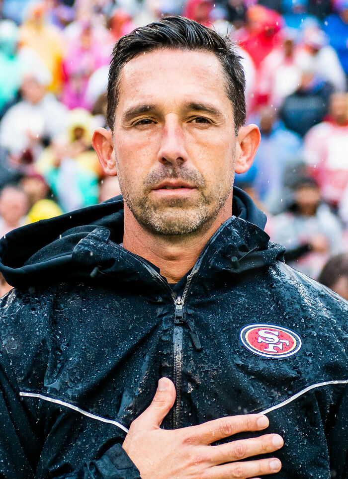 Kyle Shanahan wearing a rain jacket with the San Francisco 49ers logo, standing with hand on chest during a game.