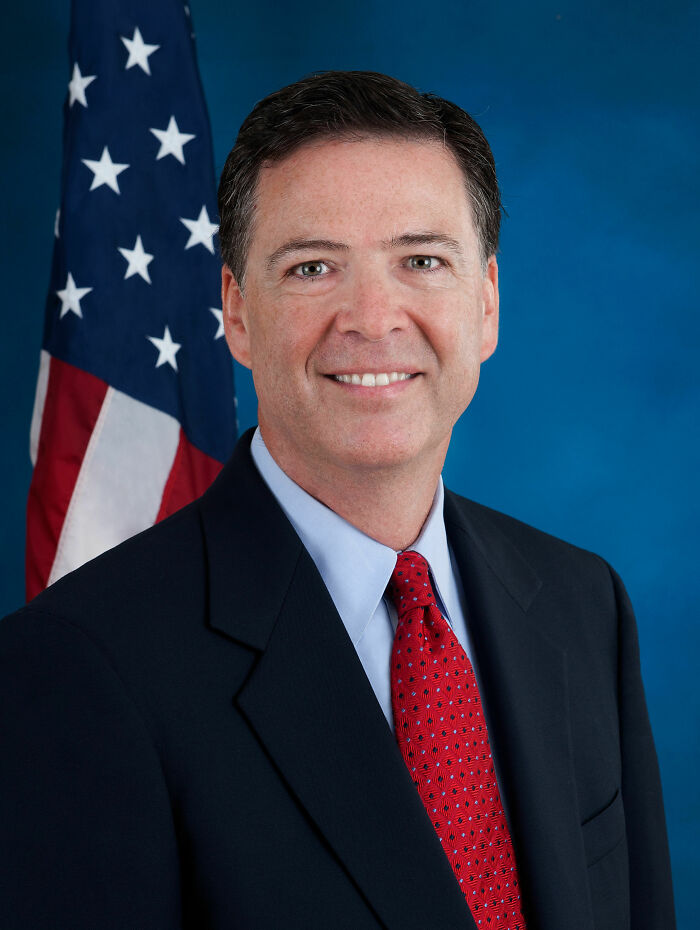James Comey smiling in a suit and red tie with an American flag backdrop, highlighting his bio and career highlights.