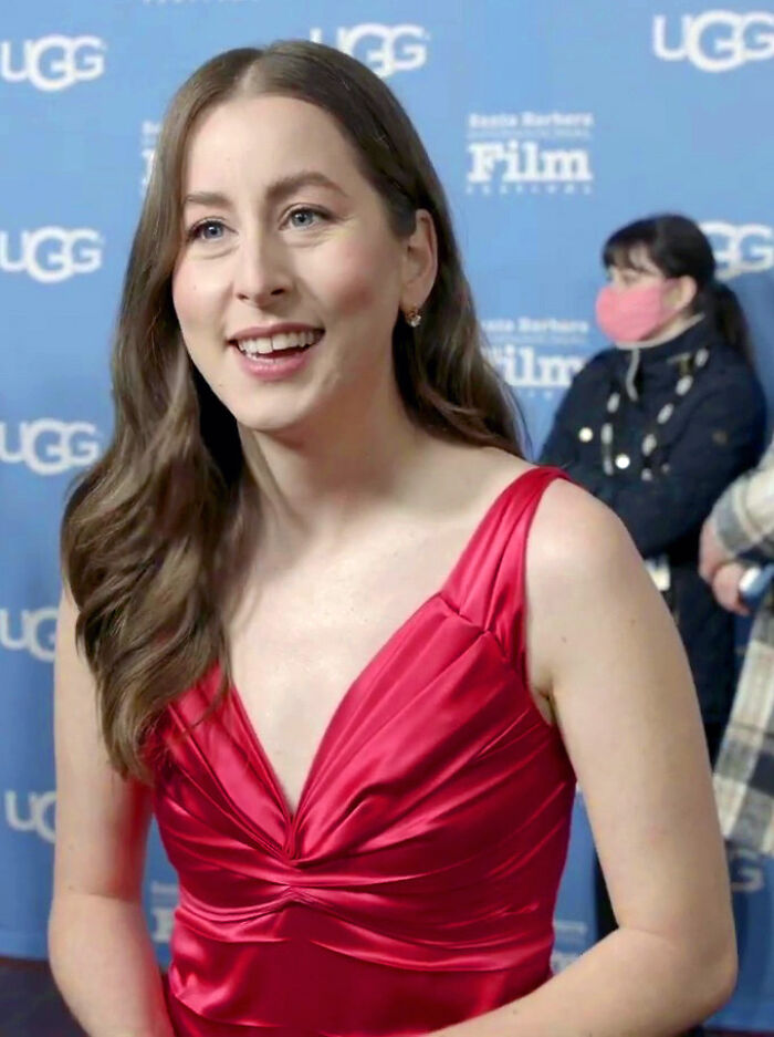 Alana Haim smiling, wearing a red dress at a film festival event with a blue background and people in the distance