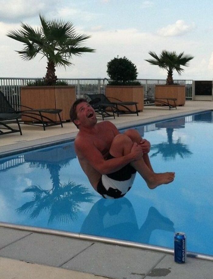 Man caught at the perfect time mid-air jump into pool with clear water reflecting palm trees and outdoor lounge chairs nearby