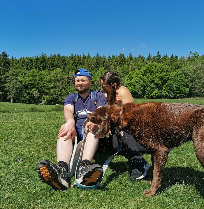 Man and woman sitting on grass with dog shaking water, funny interesting weird pic taken at the perfect time outdoors.